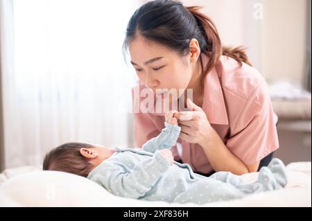 Un neonato che tiene il dito della mamma. Una giovane e premurosa mamma asiatica che mette il suo bambino a dormire in camera da letto. legame familiare, amore e tenerezza Foto Stock