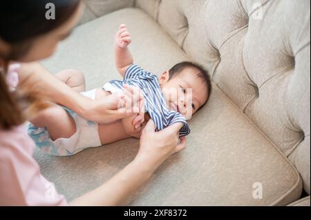 Una mamma premurosa sta cambiando i vestiti e il pannolino del suo neonato, o indossando un pannolino e un pigiama per lui sul divano del soggiorno. assistenza all'infanzia Foto Stock