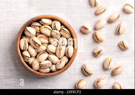 Pistacchi in conchiglie, arrostiti e salati, in ciotola di legno su lino. Croccanti frutti arrostiti di Pistacia vera, con conchiglie aperte e semi all'interno. Foto Stock