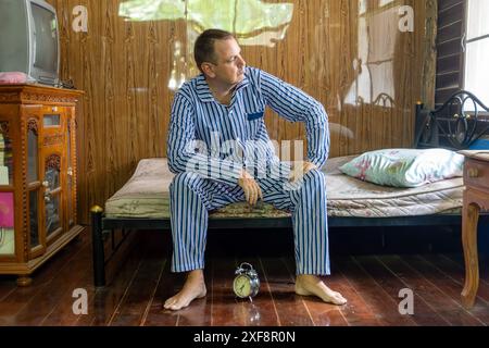 Un uomo in pigiama a righe siede su un materasso e guarda fuori dalla finestra Foto Stock