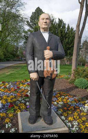 Statua di un uomo in abito scuro che tiene un violino si erge in un giardino fiorito sotto un cielo limpido, sculture a Siegen Foto Stock