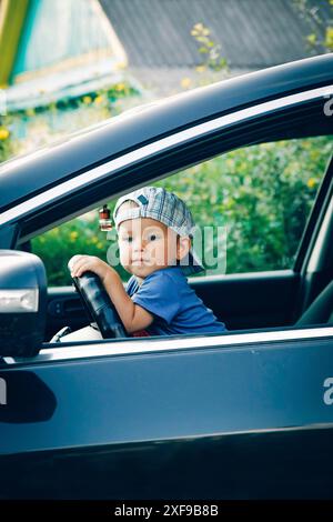 Un bambino seduto sul sedile del conducente di un'auto con una camicia e un berretto blu, sullo sfondo di una vegetazione lussureggiante. Bielorussia, Minsk Foto Stock