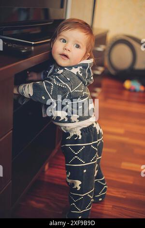 Un bambino in una felpa con cappuccio grigia con motivi di elefante bianco si trova vicino a un televisore in un accogliente soggiorno Foto Stock