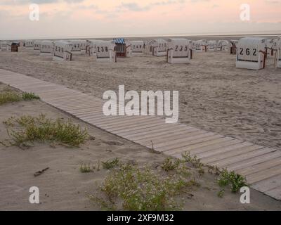 Il sentiero in legno conduce alle sdraio sulla spiaggia al tramonto, ad alcune piante sul bordo, Spiekeroog, mare del nord, germania Foto Stock