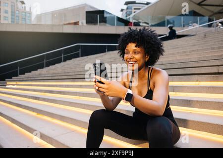 Giovane donna allegra seduta su gradini illuminati che scatta un selfie dopo una sessione di allenamento in un ambiente urbano. Foto Stock