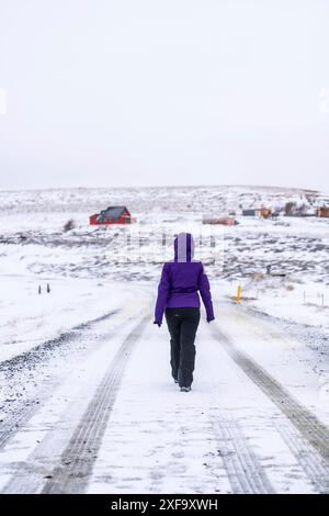 Una donna con una giacca viola cammina lungo una strada innevata. La strada è ricoperta di neve e presenta tracce di pneumatici. La donna sta camminando da sola e si sta divertendo Foto Stock