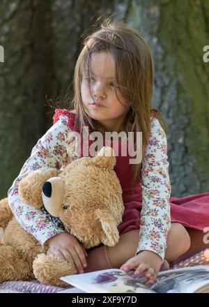 Una bambina carina di 5 anni si siede vicino a un grande albero nel parco. Tenendo in mano un grande orsacchiotto, guarda attentamente di lato. Foto Stock