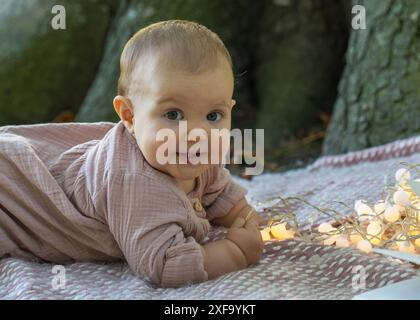 Il ritratto di una bambina affascinante in un abito beige giace sullo stomaco vicino a un grande albero, guardando la macchina fotografica. Ci sono lanterne nelle vicinanze. Il bambino io Foto Stock