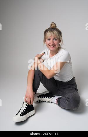 Foto a figura intera di un'adorabile donna bionda seduta sul pavimento, che indossa jeans, t-shirt bianca e scarpe da ginnastica, isolata su uno sfondo bianco. Sorride Foto Stock