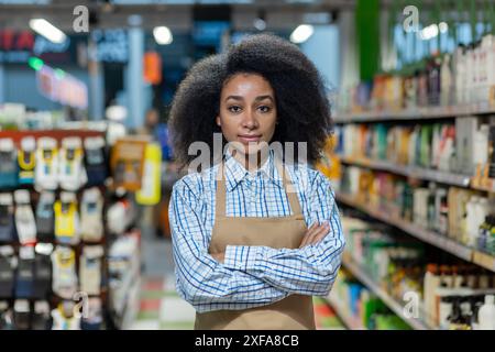 Ritratto di una donna sicura di sé che lavora al supermercato in piedi con le braccia incrociate in un corridoio di un negozio. Indossa un grembiule e una camicia a scacchi, incarnando professionalità e dedizione nel lavoro al dettaglio. Foto Stock