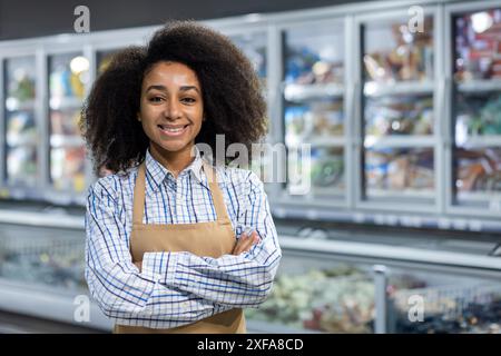 Un allegro dipendente del supermercato che indossa un grembiule e una camicia a quadri si erge con le braccia incrociate davanti alla sezione refrigerata. Trasuda cordialità e professionalità Foto Stock