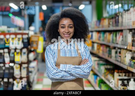 Ritratto di un dipendente sicuro del supermercato in piedi con le braccia incrociate in un corridoio. Indossando un grembiule e un sorriso cordiale, incarna professionalità e servizio clienti in un ambiente di vendita al dettaglio. Foto Stock