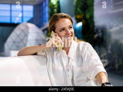 Attraente donna d'affari con una camicia bianca che parla su un telefono cellulare e sorride mentre siede in una business lounge dell'aeroporto. Foto Stock