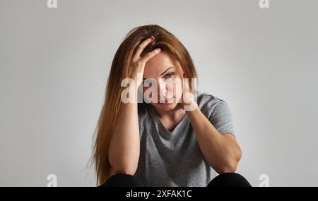Ritratto di una giovane bruna solitaria depressa con problemi mentali si trova davanti alla parete grigia e tiene la testa con la mano. Foto Stock