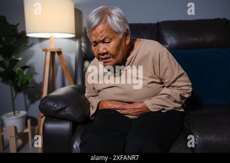 donna anziana che soffre di mal di stomaco mentre si siede su un divano nel soggiorno di notte Foto Stock