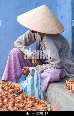 Donna anziana vietnamita in tradizionale cappello conico nella città vecchia di Hoi An, Vietnam. Anziani vietnamiti che indossano un tradizionale cappello da sole che vende per strada lui Foto Stock