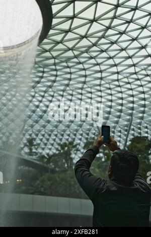 Un uomo che usa il telefono cellulare per scattare foto del vortice della pioggia a Jewel Changi, Singapore. Foto Stock