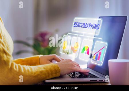 Primo piano di una persona che digita su un notebook, con la parola "BENCHMARKING" e le icone correlate che fluttuano sopra lo schermo. Ideale per gli affari, prestazioni ana Foto Stock