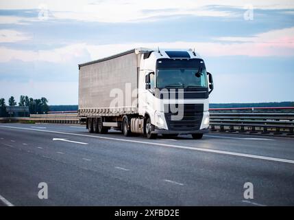 Un veicolo moderno con semirimorchio inclinabile trasporta il carico in autostrada di sera con i fari accesi contro il cielo. Concetto di trasposizione del carico Foto Stock