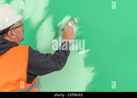 Un pittore in tuta e un casco dipinge un muro con un pennello. Copia spazio. sfondo verde Foto Stock