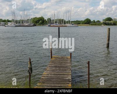 Molo in legno fatiscente sulla riva del lago con vista su diverse barche e alberi sullo sfondo. Atmosfera tranquilla sotto un cielo nuvoloso, arnis Foto Stock