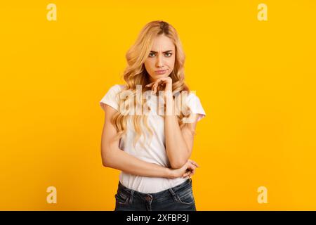 Donna bionda con la mano su Chin che pensa su sfondo giallo Foto Stock