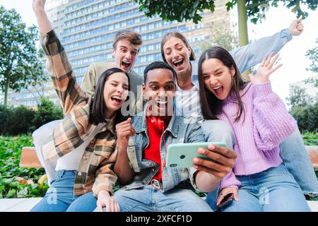 Gruppo di giovani studenti adolescenti che guardano i loro appunti su Internet usando un cellulare e celebrano con gioia il loro passaggio. Amici di successo entusiasti di applaudire con uno smartphone. Obiettivi dei compagni di classe. Foto di alta qualità Foto Stock