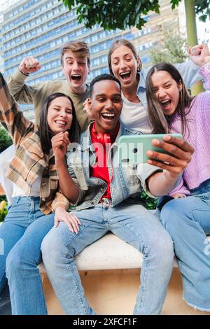 Gruppo verticale di giovani studenti adolescenti che guardano le loro note utilizzando un cellulare e celebrano con gioia il superamento. Amici di successo entusiasti di applaudire con uno smartphone. Obiettivi dei compagni di classe. Foto di alta qualità Foto Stock