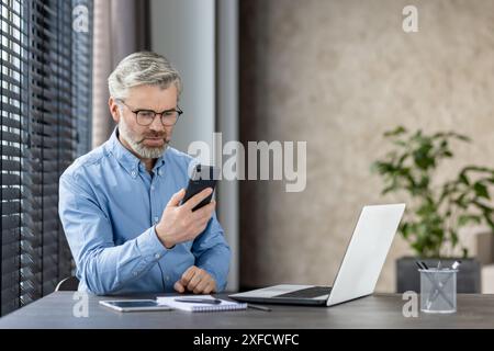 Un uomo d'affari di mezza età siede alla scrivania con un computer portatile e uno smartphone in un ufficio moderno. Sembra concentrato e premuroso, possibilmente in grado di gestire compiti o comunicazioni. Concetto di produttività aziendale Foto Stock
