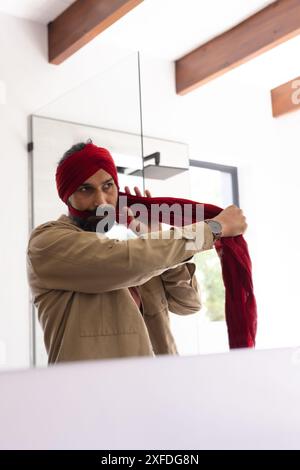 Legando il turbante rosso, uomo in piedi davanti allo specchio a casa Foto Stock