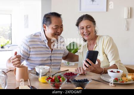 Sorridente coppia anziana che usa lo smartphone mentre gusta la colazione insieme a casa Foto Stock