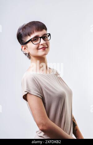 Giovane donna sicura e felice con capelli corti e occhiali alla moda sorride in un luminoso studio con sfondo bianco. Lei trasuda gioia e soddisfazione con spirito Foto Stock