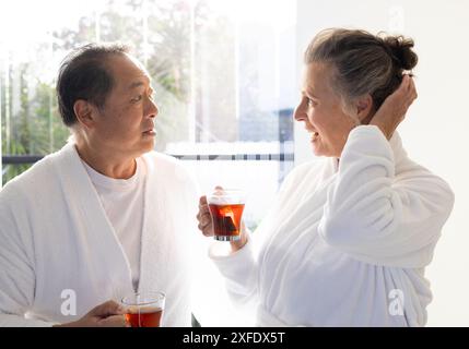 Indossando accappatoi, una coppia anziana si gode il tè e parla in una camera luminosa Foto Stock
