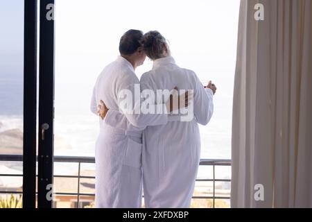 Una coppia senior in accappatoi si affaccia sul balcone e gode di vista sull'oceano Foto Stock