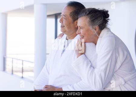 Una coppia anziana in accappatoi che si diverte in vacanza, abbracciando e sorridendo sul balcone Foto Stock