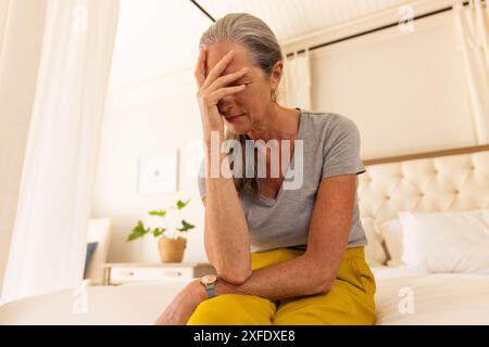 Seduta sul letto, donna matura che tiene la testa in mano, sentendosi stressata Foto Stock