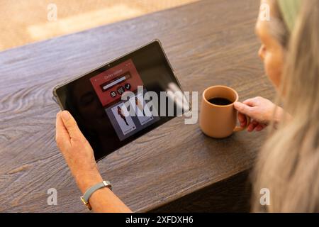 Sfogliando le opzioni di abbigliamento su tablet, donna matura che tiene una tazza di caffè a casa Foto Stock