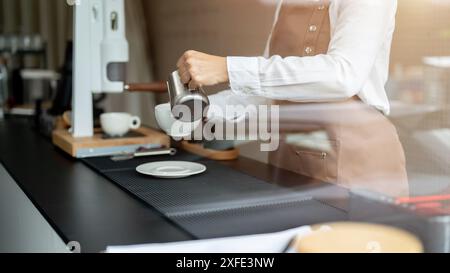 Immagine ravvicinata di una barista esperta che versa latte in una tazza da caffè, prepara gli ordini dei clienti e lavora in un bar. Foto Stock