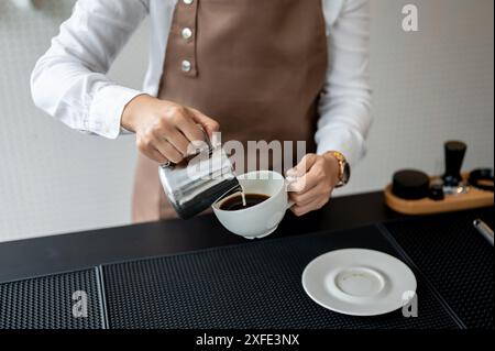 Immagine ravvicinata di una barista esperta che versa latte in una tazza da caffè, prepara gli ordini dei clienti e lavora in un bar. Foto Stock