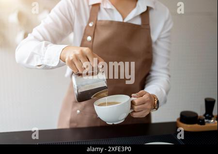 Immagine ravvicinata di una barista esperta che versa latte in una tazza da caffè, prepara gli ordini dei clienti e lavora in un bar. Foto Stock