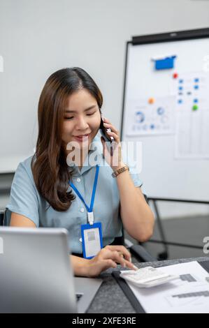 Una bella donna d'affari asiatica con esperienza o un'analista finanziaria donna sta usando una calcolatrice mentre parla al telefono con il suo cliente, lavorando su Foto Stock
