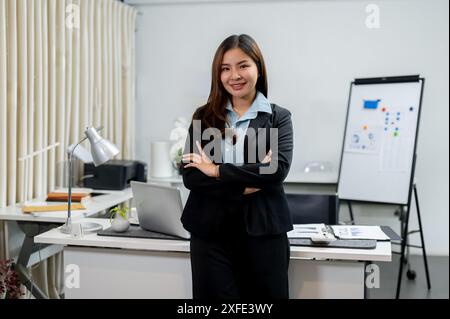 Una donna d'affari asiatica sicura di sé con un costume da lavoro è in piedi con le braccia incrociate in ufficio, sorridendo alla telecamera. concetto di uomini d'affari Foto Stock