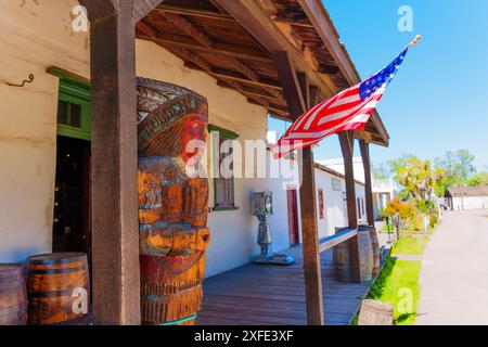 San Diego, California - 16 aprile 2024: Statua in legno dei nativi americani in piedi su un portico con una bandiera americana appesa sopra. Vetrina e botti Foto Stock