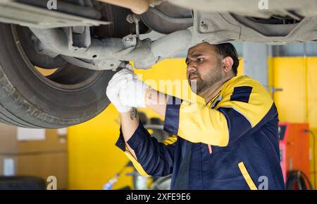 Uomo in uniforme che lavora sotto un'auto sollevata su un rack di sollevamento idraulico, in un'officina di riparazione automobilistica. La serie di utensili per il fissaggio meccanico Foto Stock