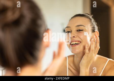 Applicazione di crema viso, giovane donna sorridente e che guarda lo specchio in bagno Foto Stock