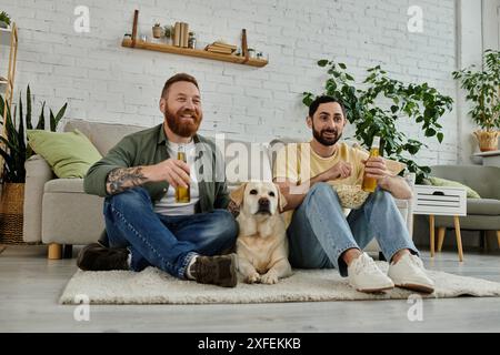 Due uomini con la barba si rilassano su un divano, guardando una partita sportiva con il loro cane Labrador nel soggiorno. Foto Stock