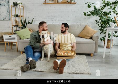 Due uomini con la barba si rilassano su un divano con una scatola di pizza, condividendo una fetta mentre un simpatico Labrador guarda. Foto Stock