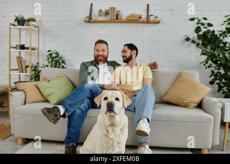 Due uomini con la barba si rilassano su un divano con il loro Labrador in un accogliente soggiorno. Foto Stock