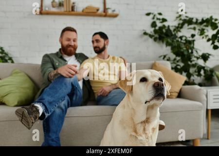 Due uomini barbuti, una coppia gay, si siedono su un divano con il loro leale labrador, godendosi un momento intimo insieme nel loro salotto. Foto Stock