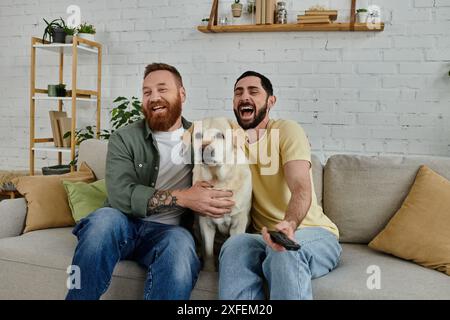 Una coppia gay barbuta siede su un divano, coccolando il loro felice labrador in un ambiente caldo e accogliente. Foto Stock